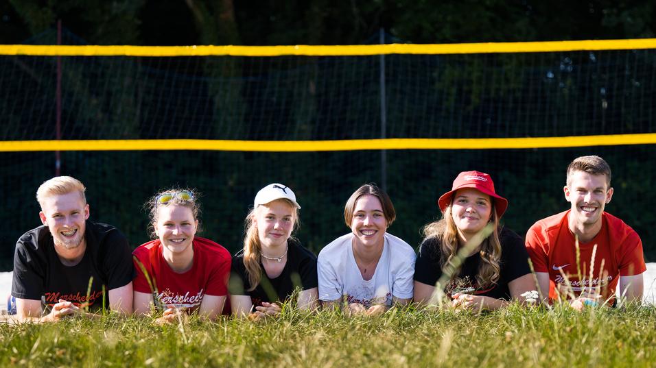 Sechs Personen liegen im Gras vor einem Beachvolleyballfeld mit gelben Netzen und Bäumen im Hintergrund.