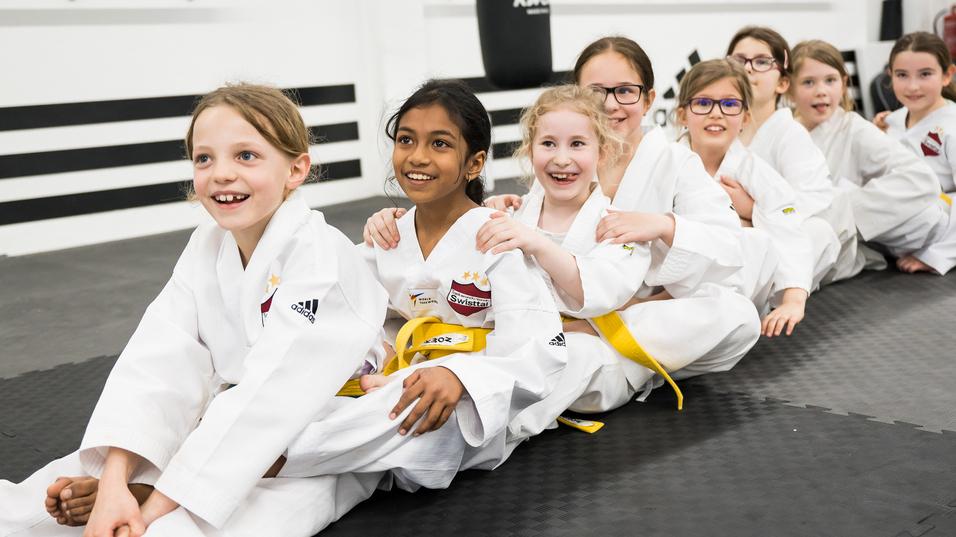 Gruppe von lächelnden Mädchen in weißen Karate-Anzügen sitzt auf einer Matte in einem Trainingsraum.